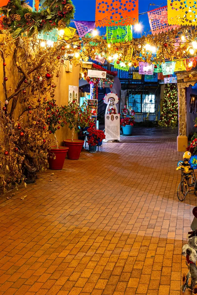 A brick walkway decorated with colorful papel picado banners and string lights, leading toward a courtyard at dusk.