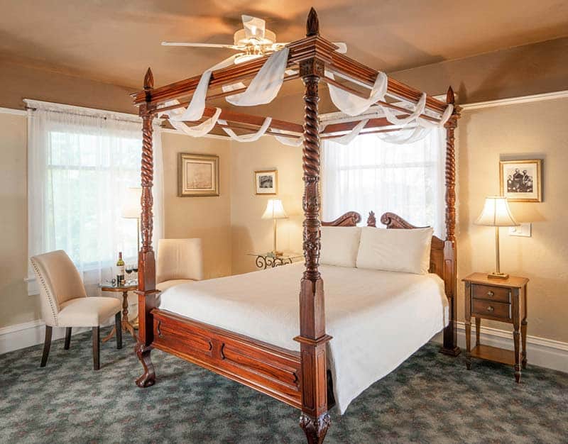A bedroom with a wooden four-poster bed, white bedding, draped fabric, beige chairs, and two lamps.