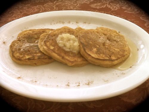 Three pancakes served on a white oval plate with syrup and a dollop of topping, set on a textured orange tablecloth.