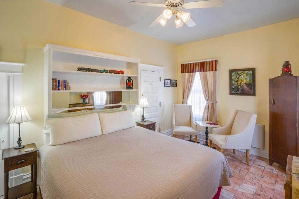 A furnished bedroom with a bed, two armchairs, a wooden cabinet, ceiling fan, and yellow walls.