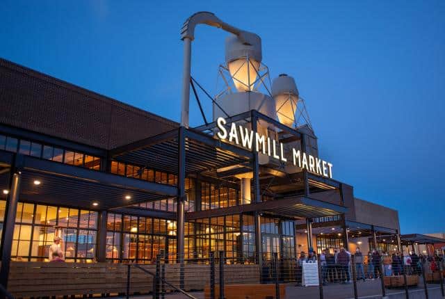 Sawmill Market exterior at dusk, featuring industrial grain silos, large windows, and a lit sign against a blue sky.