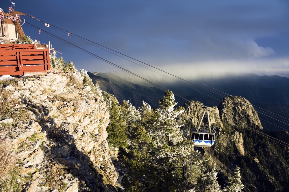 A cable car moves through the air near a rocky, tree-covered mountain peak under a cloudy sky.