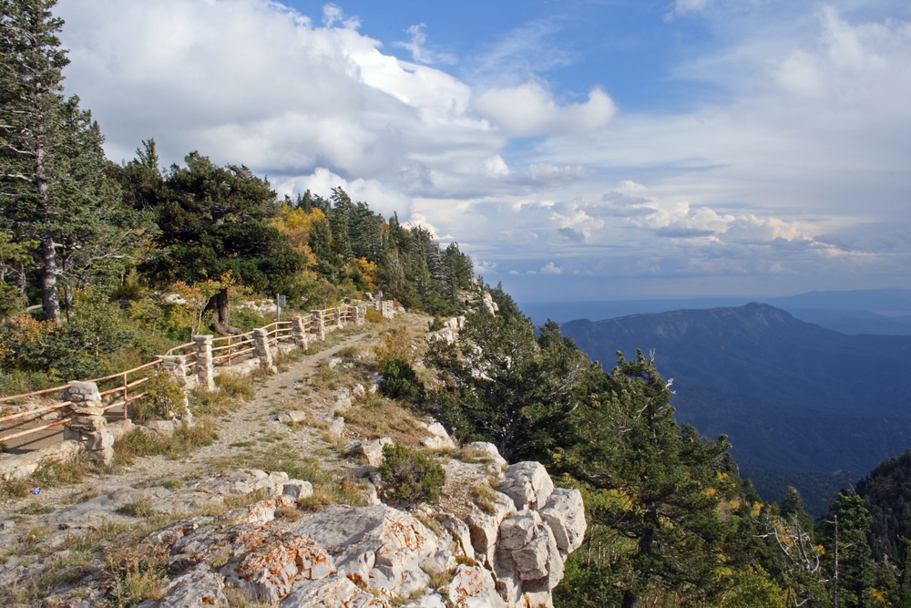 A dirt path with a rustic wooden fence runs along the edge of a mountain cliff overlooking a valley under a cloudy sky.