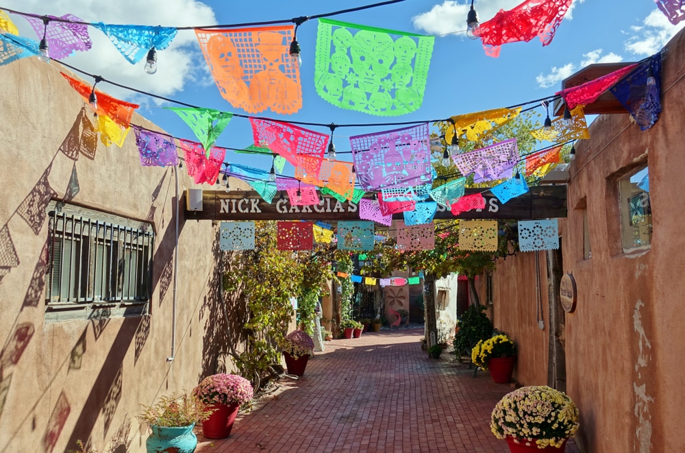 A brick courtyard path between two adobe walls is decorated overhead with colorful cutout papel picado banners.