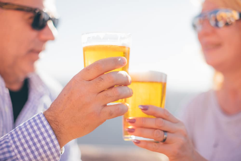 Two people wearing sunglasses toast each other with glasses of golden beer, viewed close up.