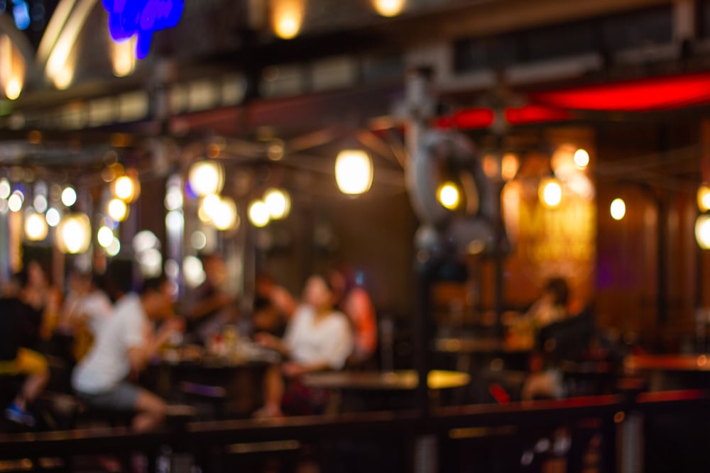 A blurry, out-of-focus night scene of people sitting at tables in a dimly lit restaurant or outdoor cafe.