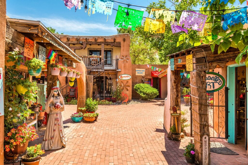 A sunlit, brick-paved courtyard decorated with colorful paper banners, potted plants, and adobe-style architecture.