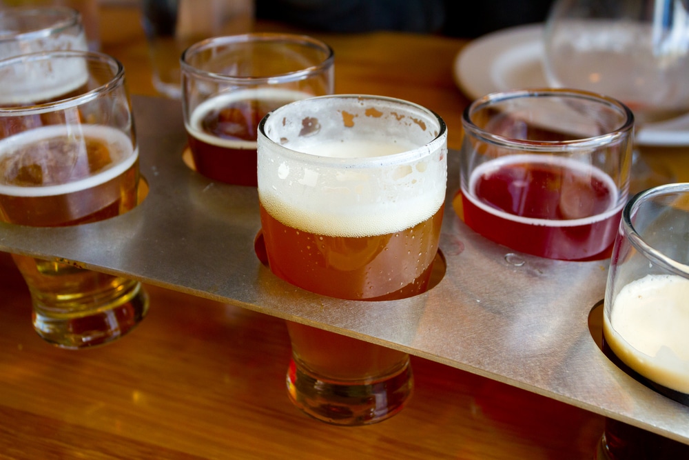 A beer flight with several small glasses of different colored beers set in a metal paddle on a wooden table.