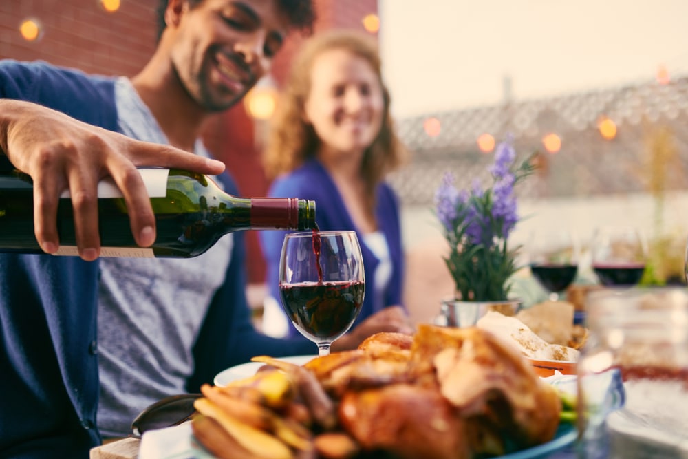 A smiling man pours red wine into a glass while a woman sits opposite him at a dinner table.