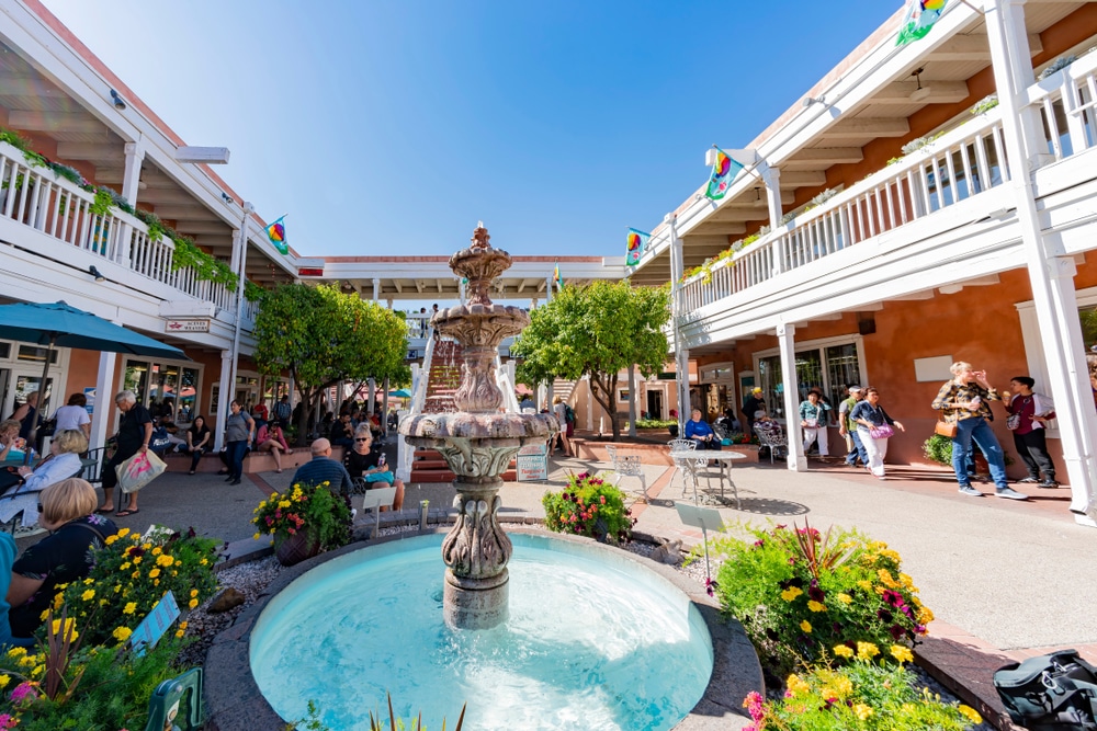 An outdoor courtyard with a tiered stone fountain, surrounding buildings with balconies, and people sitting at tables.