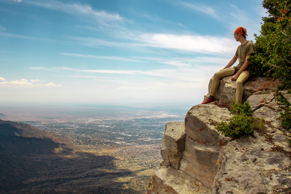 A person with red hair sits on a rocky cliff edge, looking out over a wide landscape under a blue sky.