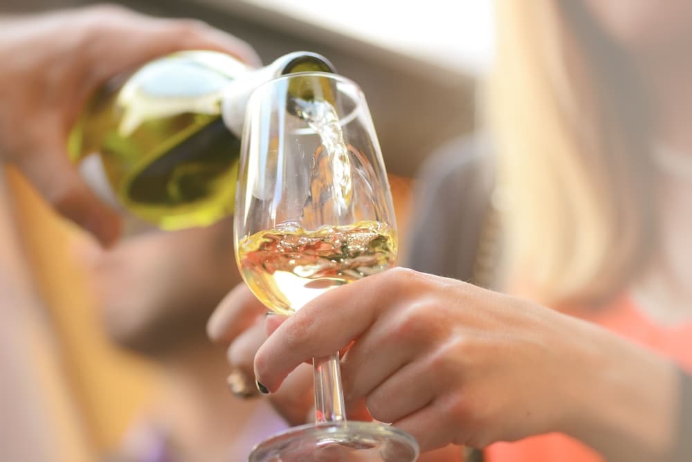 A person pours white wine from a bottle into a glass held by another individual, with soft, out-of-focus lighting.