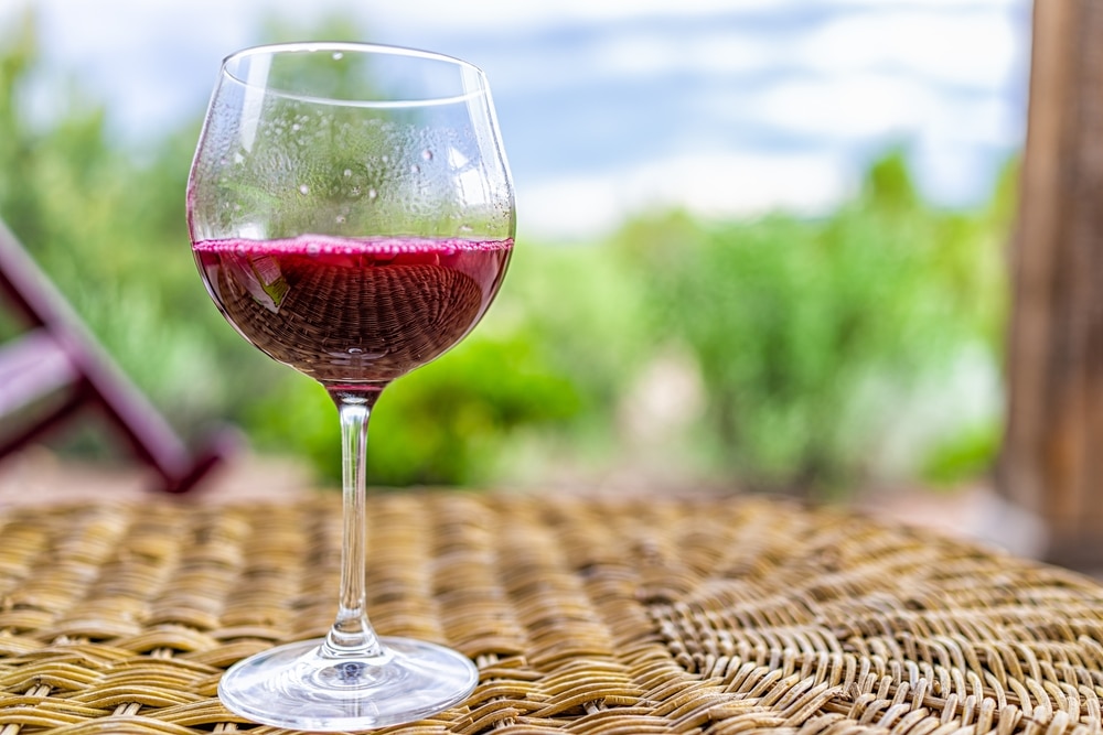A glass of red wine sits on a wicker table with a blurred outdoor background.
