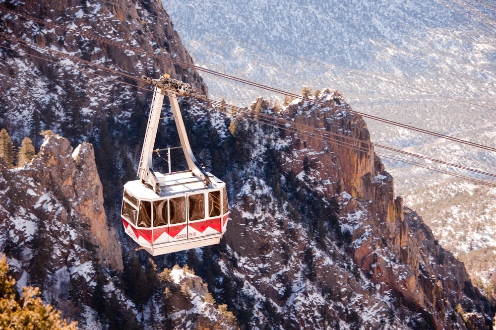 A white and red aerial tram car traverses a cable over snow-dusted, craggy mountain cliffs.