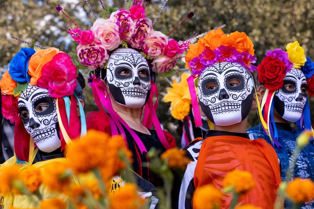 People in sugar skull face paint and floral headpieces pose behind marigold flowers, celebrating Day of the Dead.