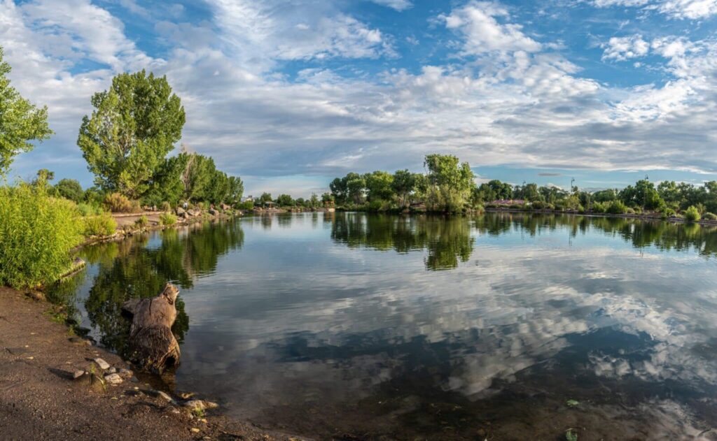 A calm lake reflects a cloudy blue sky, surrounded by lush green trees and a dirt shoreline.