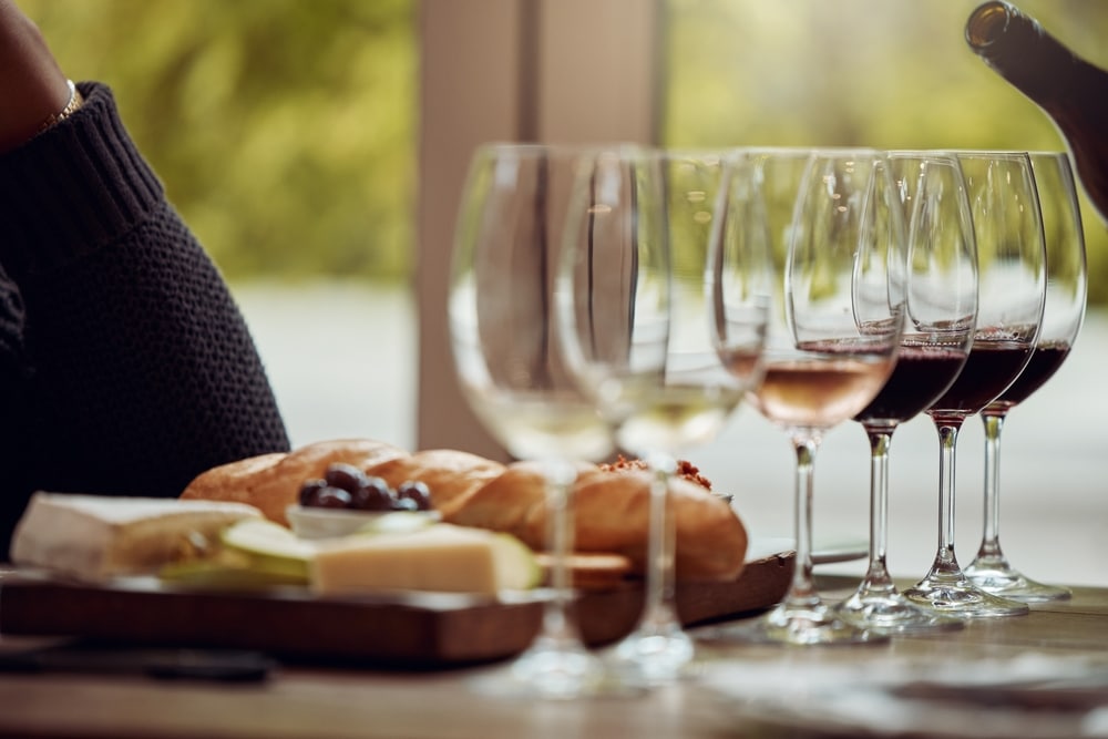 A row of wine glasses filled with different wines stands on a table beside a wooden board with bread and cheese.
