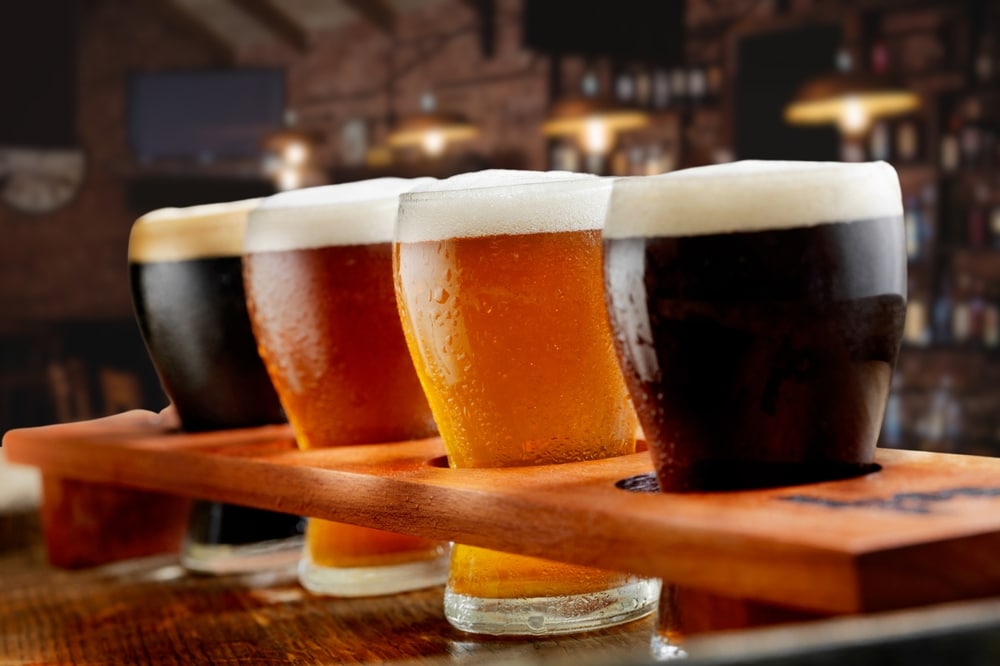 A beer flight with four different glasses of beer sits on a wooden board in a dimly lit bar setting.