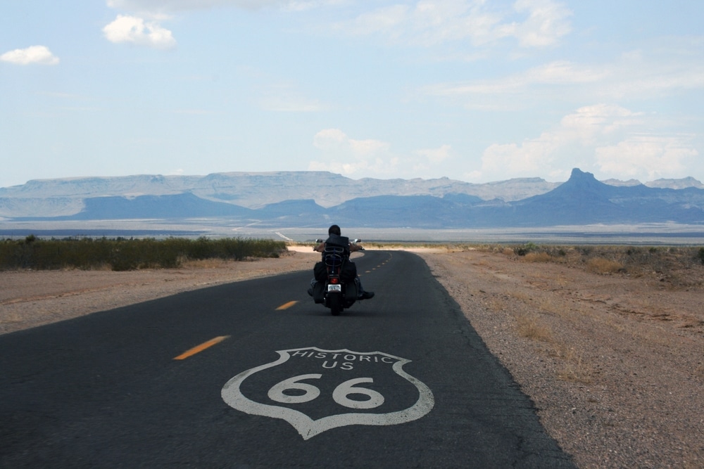 A motorcyclist rides down a desert road marked with a "Historic US 66" shield, set against distant mountains.