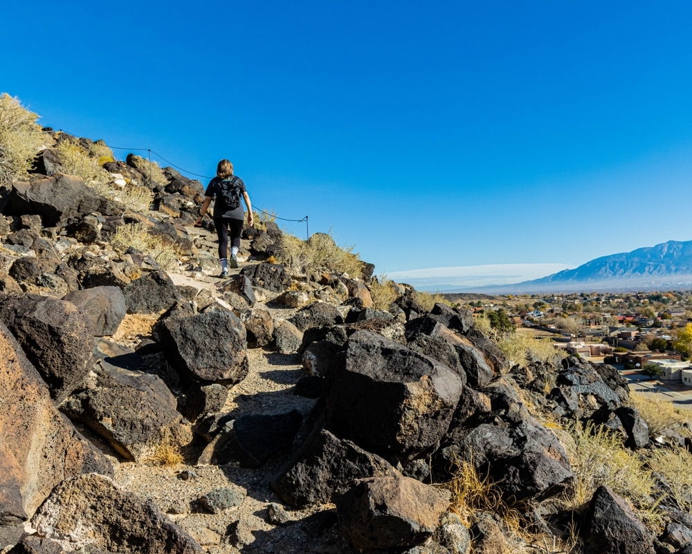 A hiker with a backpack climbs a rocky trail overlooking a valley under a clear blue sky.