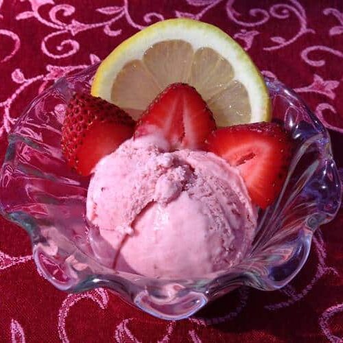 A glass bowl holds strawberry ice cream topped with sliced strawberries and a lemon wedge on a patterned red tablecloth.