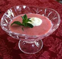 A crystal bowl holds strawberry soup topped with a dollop of cream and a mint sprig on a red tablecloth.