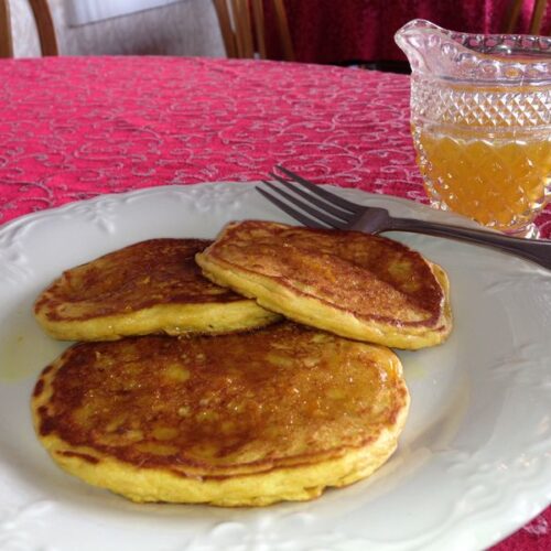 Three golden pancakes are served on a white plate with a fork and a glass pitcher of syrup on a red patterned tablecloth.