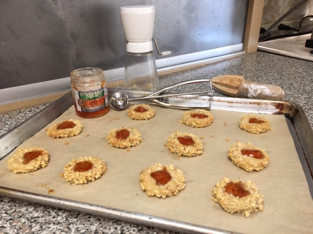 A baking sheet of thumbprint cookies with jam centers sits on a counter with a jar of jam and a cookie scoop.